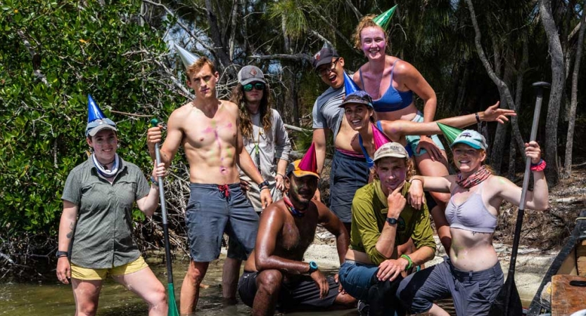 A group of people stand in shallow water and pose for a photo. Some are holding paddles, and some are wearing party hats.